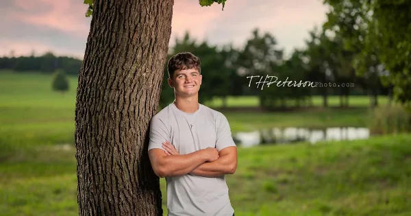 A young man in a grey t-shirt leaning against a tree trunk in a grassy park with a pond in the background.