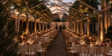 Outdoor event reception under a clear-frame tent with string lights, round tables, chairs, and floral centerpieces at dusk.