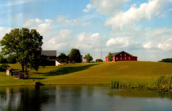 A wide shot of a farm property featuring a red barn, a pond in the foreground, and green rolling hills under a blue sky.