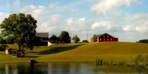 A wide shot of a farm property featuring a red barn, a pond in the foreground, and green rolling hills under a blue sky.