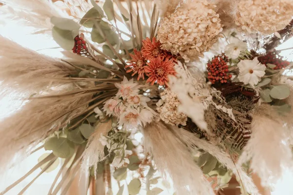 A close-up of a wedding floral arrangement featuring pampas grass, dried hydrangeas, and orange and white flowers.