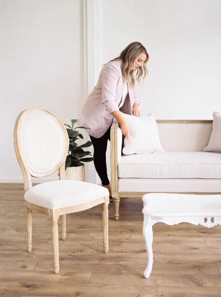 A woman arranging a pillow on a cream-colored sofa next to a vintage wooden chair and a white table in a bright room.