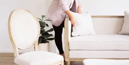 A woman arranging a pillow on a cream-colored sofa next to a vintage wooden chair and a white table in a bright room.