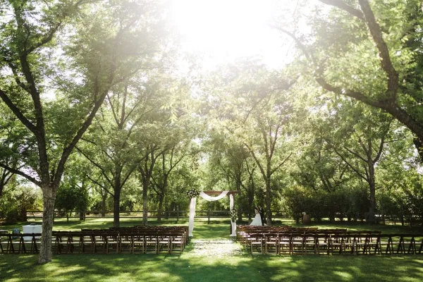 An outdoor wedding ceremony setup with rows of chairs and a floral arch under a canopy of green trees.