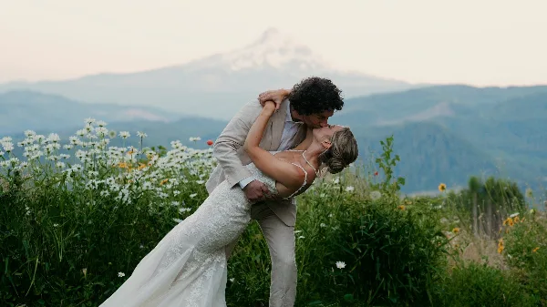 A cinematic photograph of a bride and groom kissing in a field of wildflowers with a mountain in the background.
