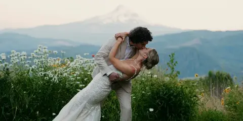 A cinematic photograph of a bride and groom kissing in a field of wildflowers with a mountain in the background.
