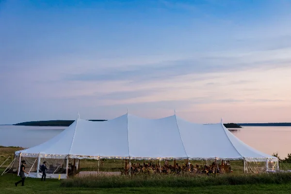 A large white event tent set up on a grassy waterfront area with guests seated inside for a reception.