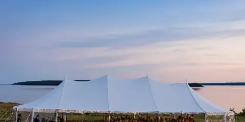 A large white event tent set up on a grassy waterfront area with guests seated inside for a reception.