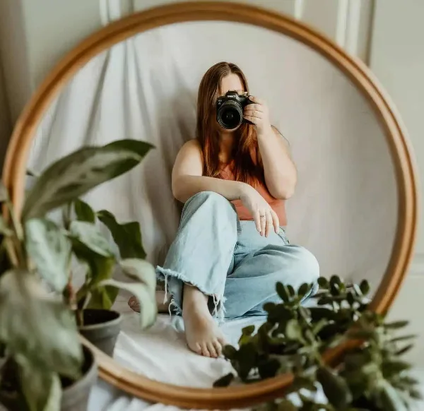 A woman holding a DSLR camera, sitting in front of a round mirror with houseplants in the foreground.