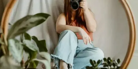 A woman holding a DSLR camera, sitting in front of a round mirror with houseplants in the foreground.