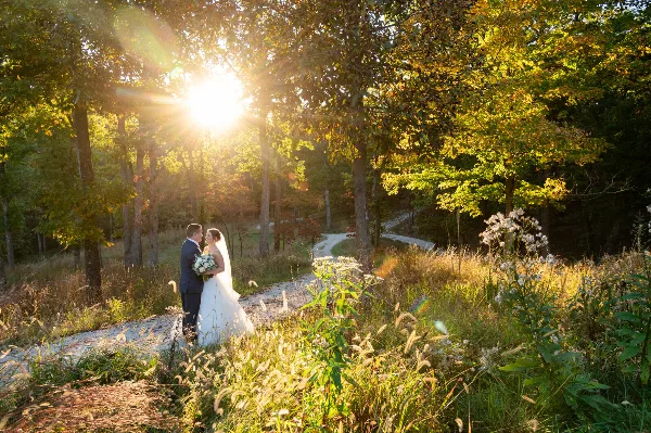 A bride and groom standing on a gravel path in a wooded area during golden hour, with the sun shining through the trees.