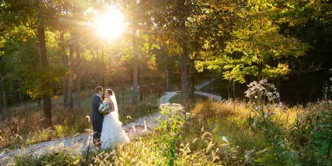 A bride and groom standing on a gravel path in a wooded area during golden hour, with the sun shining through the trees.