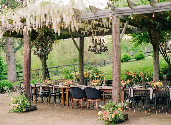 An outdoor wedding reception setup featuring a long wooden table, floral arrangements, and a pergola with hanging lights.