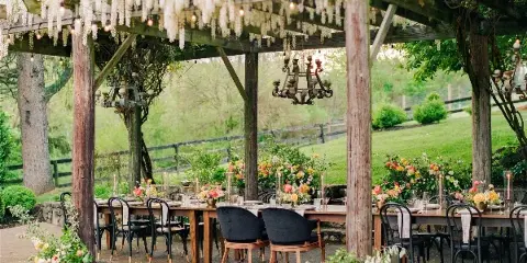 An outdoor wedding reception setup featuring a long wooden table, floral arrangements, and a pergola with hanging lights.