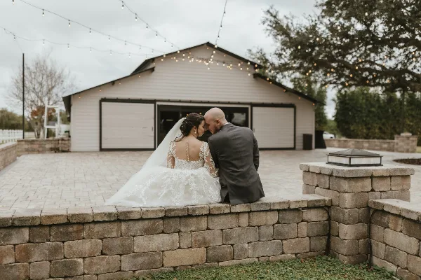 Wedding couple seated on a stone terrace in front of a rustic barn with string lights
