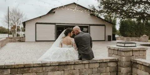 Wedding couple seated on a stone terrace in front of a rustic barn with string lights