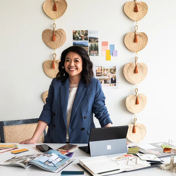 A smiling woman in a blue blazer stands behind a desk covered with wedding planning materials, color swatches, and design portfolios.