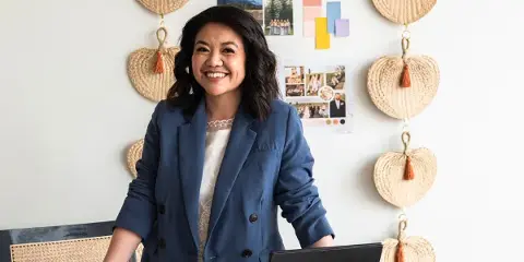 A smiling woman in a blue blazer stands behind a desk covered with wedding planning materials, color swatches, and design portfolios.