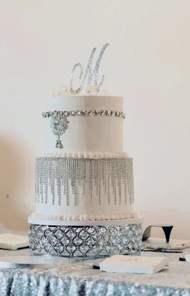 A two-tier white wedding cake decorated with silver crystal fringe, a jeweled brooch, and a sparkling monogram topper.