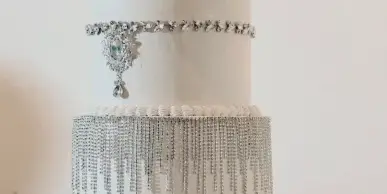 A two-tier white wedding cake decorated with silver crystal fringe, a jeweled brooch, and a sparkling monogram topper.
