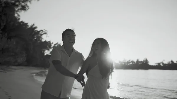 Black-and-white wedding film still of a smiling couple holding hands on a tropical beach at sunset.