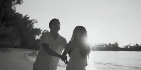 Black-and-white wedding film still of a smiling couple holding hands on a tropical beach at sunset.
