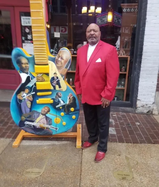 A man in a red suit standing next to a guitar sculpture featuring portraits of musicians.