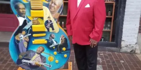 A man in a red suit standing next to a guitar sculpture featuring portraits of musicians.