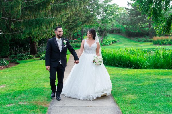 A bride and groom holding hands and walking along a paved path in a lush green garden setting.
