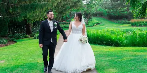 A bride and groom holding hands and walking along a paved path in a lush green garden setting.