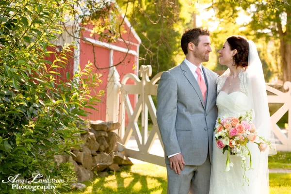 A bride and groom standing together outdoors near a red barn and white fence, with the bride holding a bouquet of pink and white flowers.
