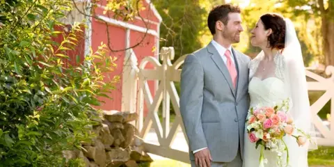 A bride and groom standing together outdoors near a red barn and white fence, with the bride holding a bouquet of pink and white flowers.