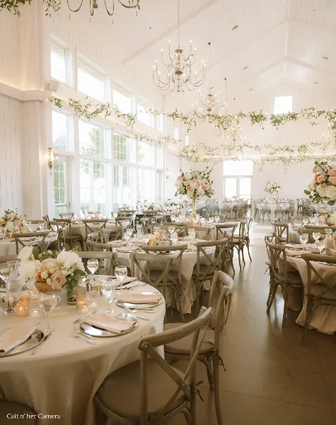 Interior view of a white barn-style wedding reception hall with round tables, chandeliers, and large windows.