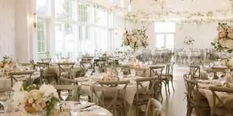 Interior view of a white barn-style wedding reception hall with round tables, chandeliers, and large windows.
