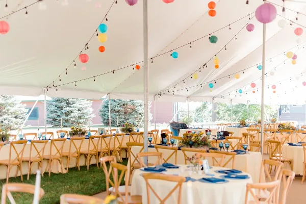 A wedding reception setup inside a white tent featuring tables with blue napkins, floral centerpieces, and colorful paper lanterns hanging from the ceiling.