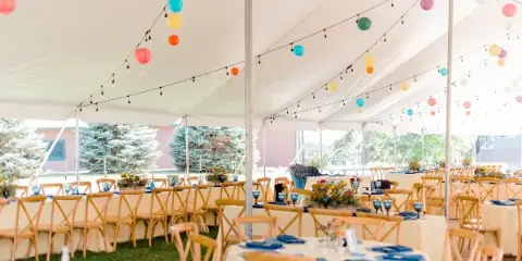 A wedding reception setup inside a white tent featuring tables with blue napkins, floral centerpieces, and colorful paper lanterns hanging from the ceiling.