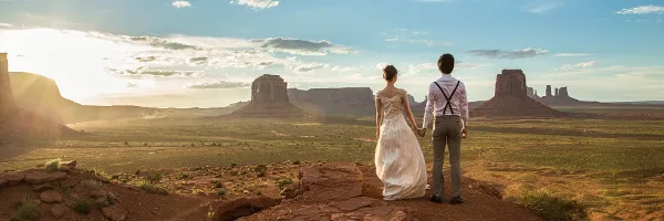 A bride and groom standing on a red rock ledge overlooking Monument Valley at sunset.