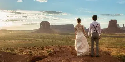 A bride and groom standing on a red rock ledge overlooking Monument Valley at sunset.