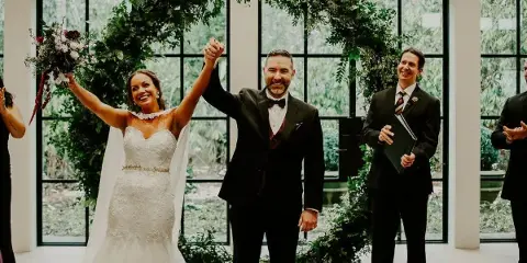 A bride and groom with raised hands celebrating during a wedding ceremony, with an officiant standing to the side.