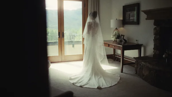 A bride in a white wedding gown and veil stands in a sunlit room with a view of mountains.