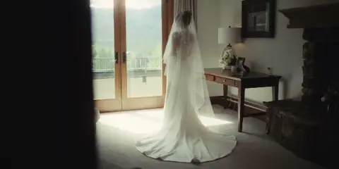 A bride in a white wedding gown and veil stands in a sunlit room with a view of mountains.