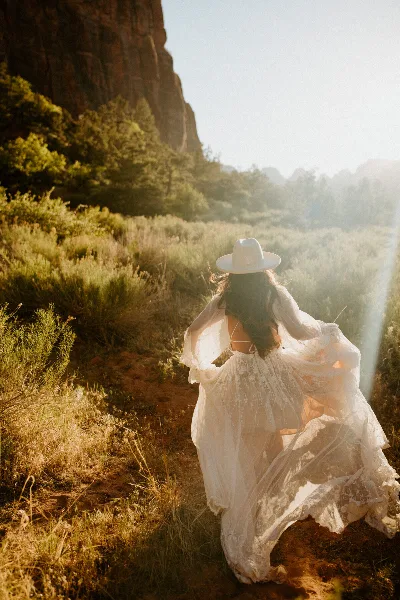 A bride in a flowing white dress and white hat walking away from the camera on a dirt path in a mountainous landscape with golden hour lighting.