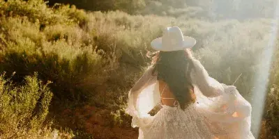 A bride in a flowing white dress and white hat walking away from the camera on a dirt path in a mountainous landscape with golden hour lighting.