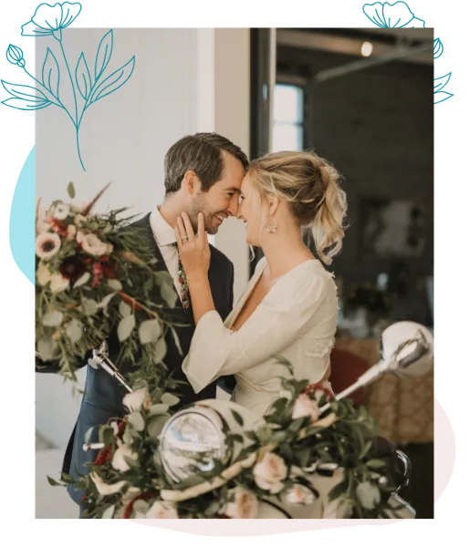 A bride and groom posing with a large, lush floral arrangement featuring white and pink flowers and greenery.