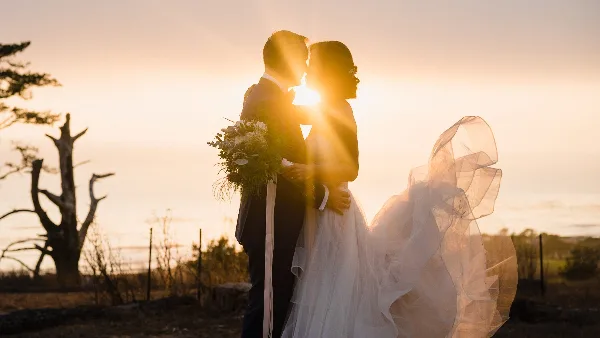 A bride and groom embracing in a field at sunset, with the bride holding a bouquet and her veil blowing in the wind.
