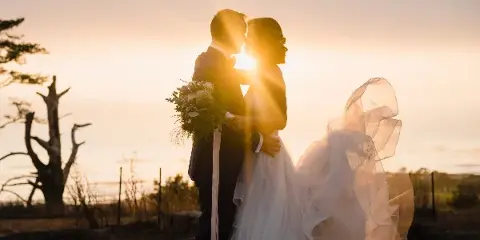 A bride and groom embracing in a field at sunset, with the bride holding a bouquet and her veil blowing in the wind.