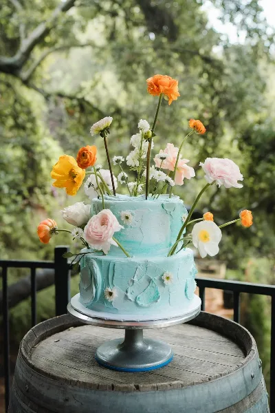 A two-tier light blue textured cake decorated with fresh orange, yellow, and white flowers on a silver stand.