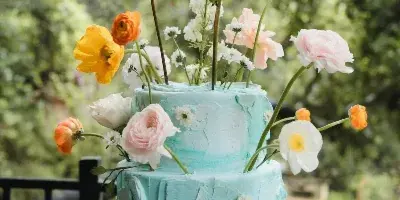 A two-tier light blue textured cake decorated with fresh orange, yellow, and white flowers on a silver stand.