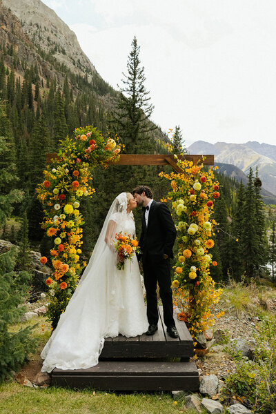 A wedding couple kissing under a wooden arch decorated with vibrant orange and yellow flowers in a mountainous outdoor setting.