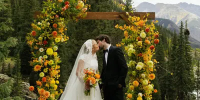 A wedding couple kissing under a wooden arch decorated with vibrant orange and yellow flowers in a mountainous outdoor setting.